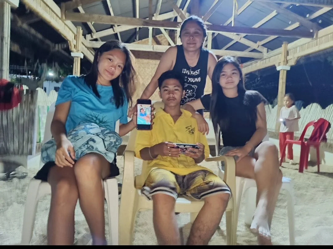 Four friends, three women and one man, are at an indoor beach area with sand and a wooden roof structure. The man is sitting in a yellow chair holding a smartphone, while the women are sitting or standing around him, smiling. One woman is holding up 