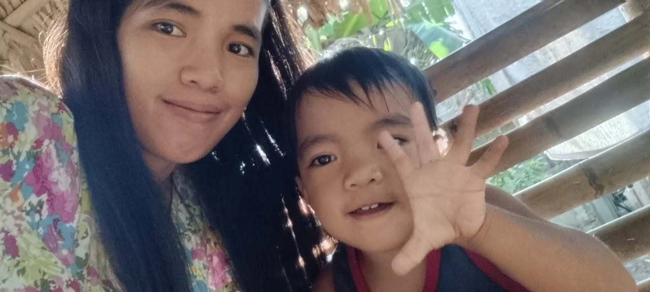 Young girl with long dark hair and a colorful floral shirt smiling while taking a selfie with a young boy. The boy has short dark hair, is holding up a hand near his face, and is wearing a dark shirt with red trim. They are outdoors with bamboo and g