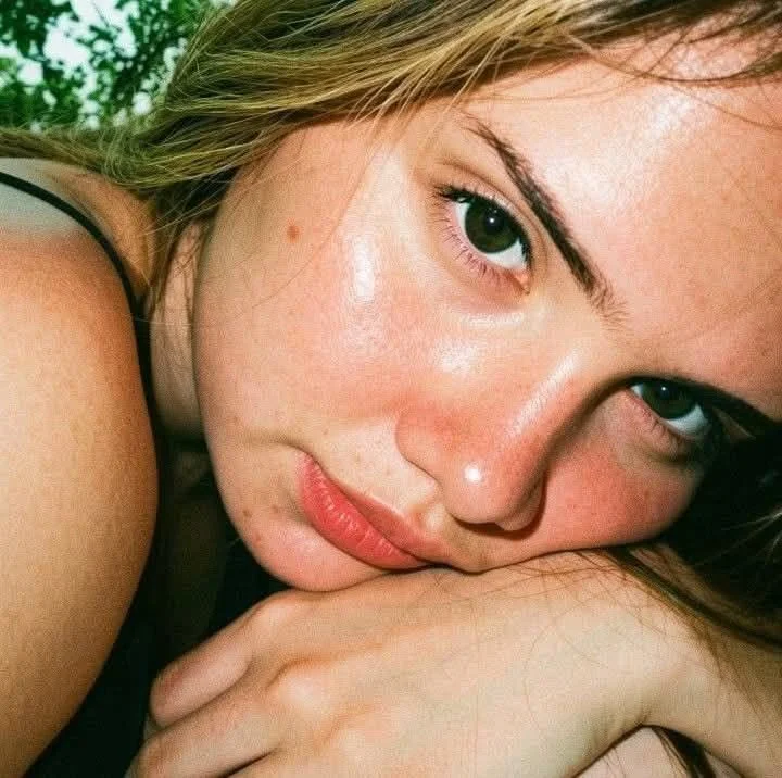 Close-up of a young woman with blue eyes resting her face on her hand, looking at the camera with a soft expression, with greenery in the background.