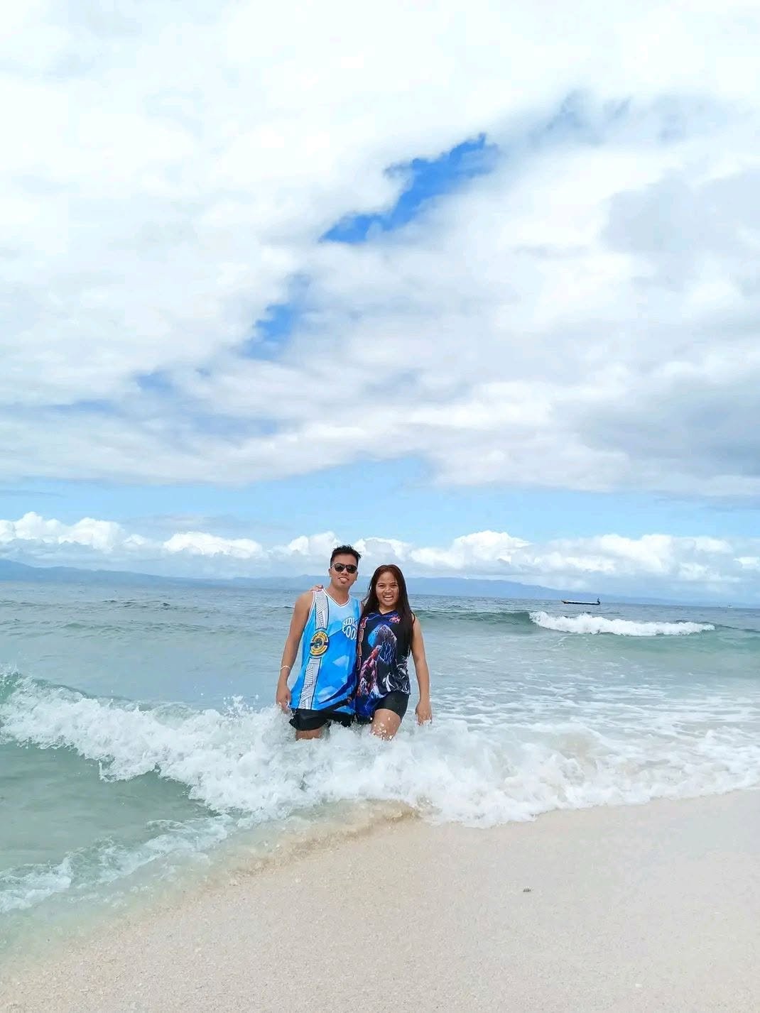 Two people standing in the ocean waves on a beach, smiling, with clouds and a boat in the background.