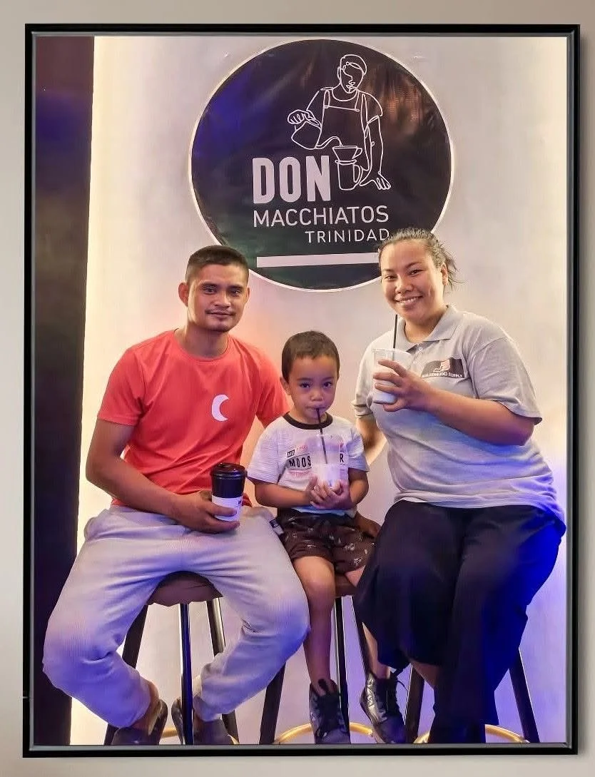 Three people, a man, a woman, and a young boy, sitting on stools in front of a sign for Don Macchiatos Trinidad, holding drinks with straws.