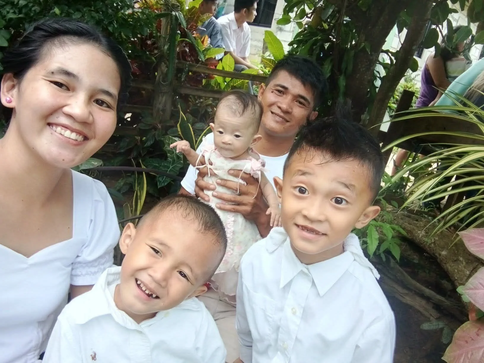 A family of five posing for a photo outdoors among lush green plants. A woman and two young boys are in the foreground, with a man holding a young girl in the background. Everyone is smiling, and the children are dressed in white shirts.