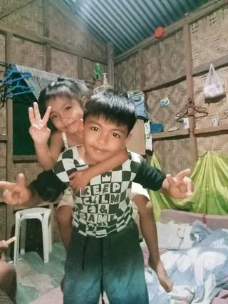 Two young boys smiling and making peace signs in a rustic room with wooden walls and a tin roof. One boy is sitting on a white stool, and the other is standing in front.