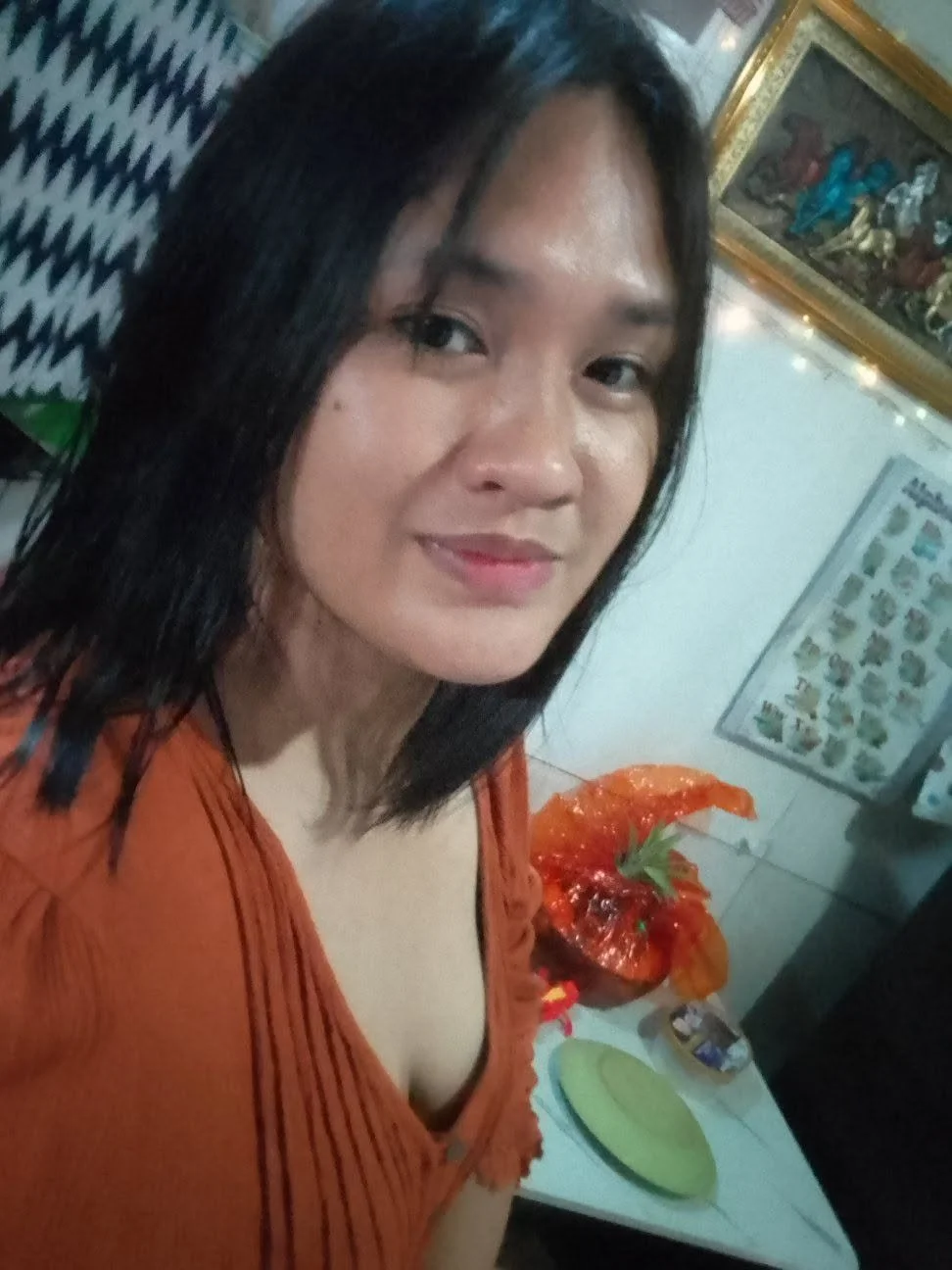A woman with shoulder-length black hair taking a selfie indoors. She is wearing an orange top and is smiling.