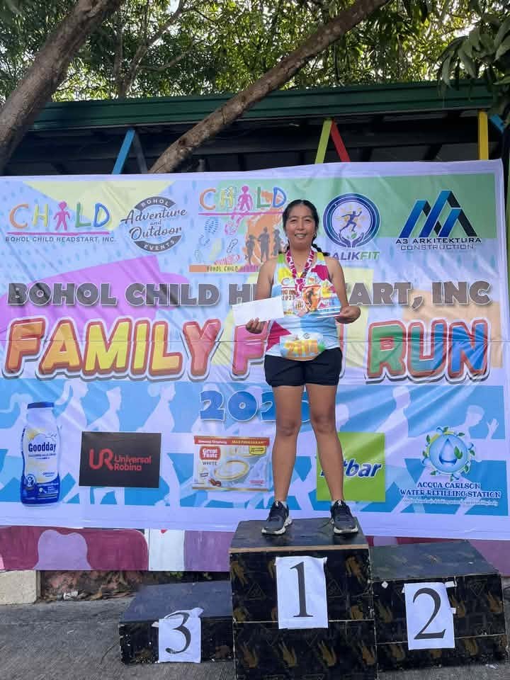A woman standing on a first place podium at a family fun run event, holding a certificate and medal, with a banner in the background promoting the Bohol Child Heart Inc. event.