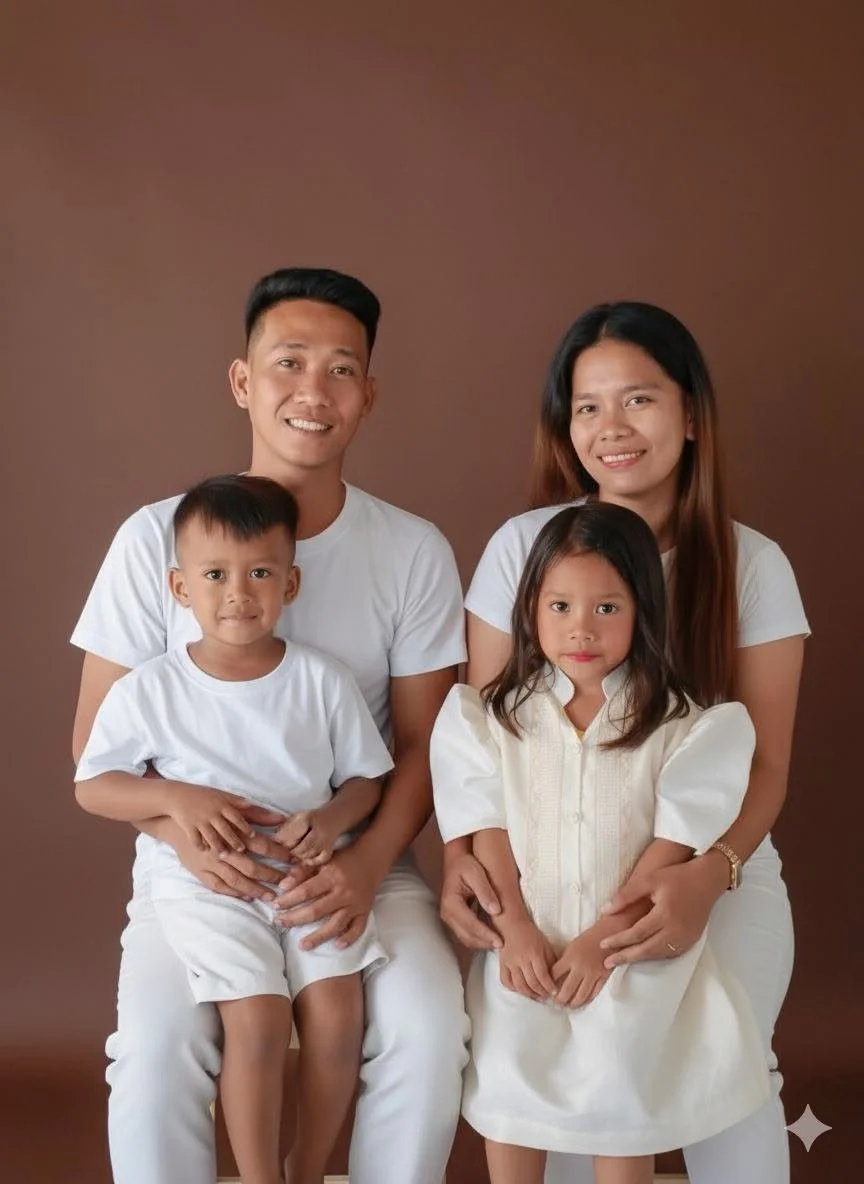 A family of four posing for a photo against a brown background. The father and mother are sitting, with the father holding a young boy on his lap and the mother holding a young girl on her lap. All are wearing white clothing.