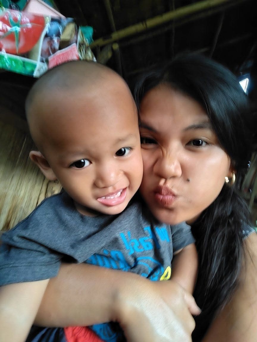 A young woman and a small boy making silly faces close together, indoors with a wooden wall background.