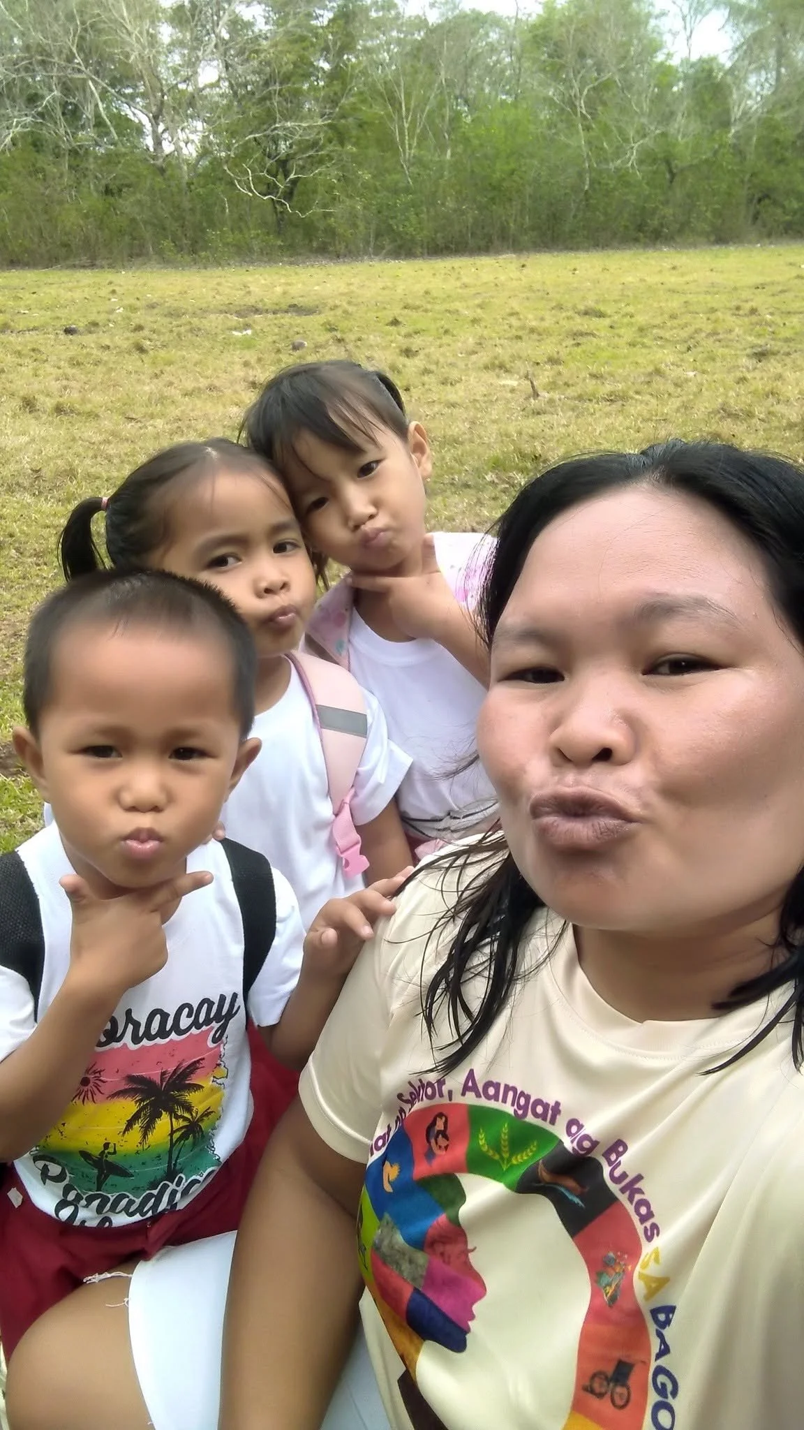 A woman and three children taking a selfie outdoors on a grassy field with trees in the background. The children and woman are making playful expressions.