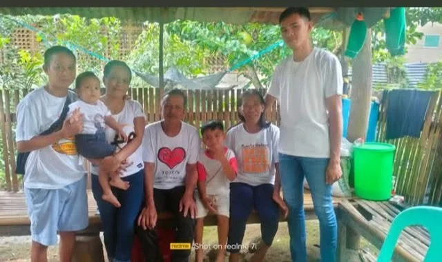 Group of seven people, including children and adults, posing together in a rustic outdoor setting with wooden fencing and greenery in the background.