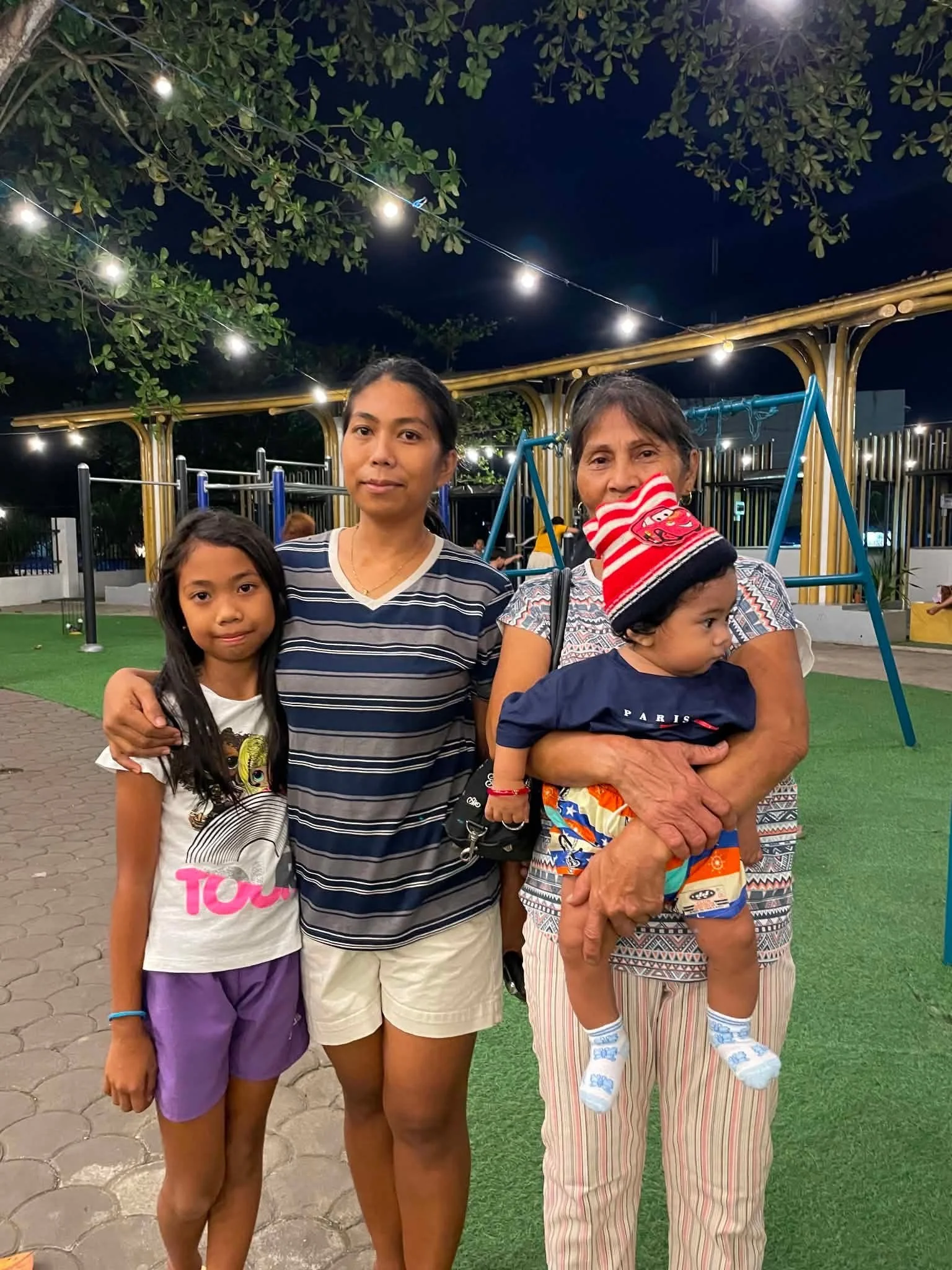 A group of people at a park at night, with two women and two children, one woman holding a young boy. The park has string lights, trees, a swing set, and a fence in the background.
