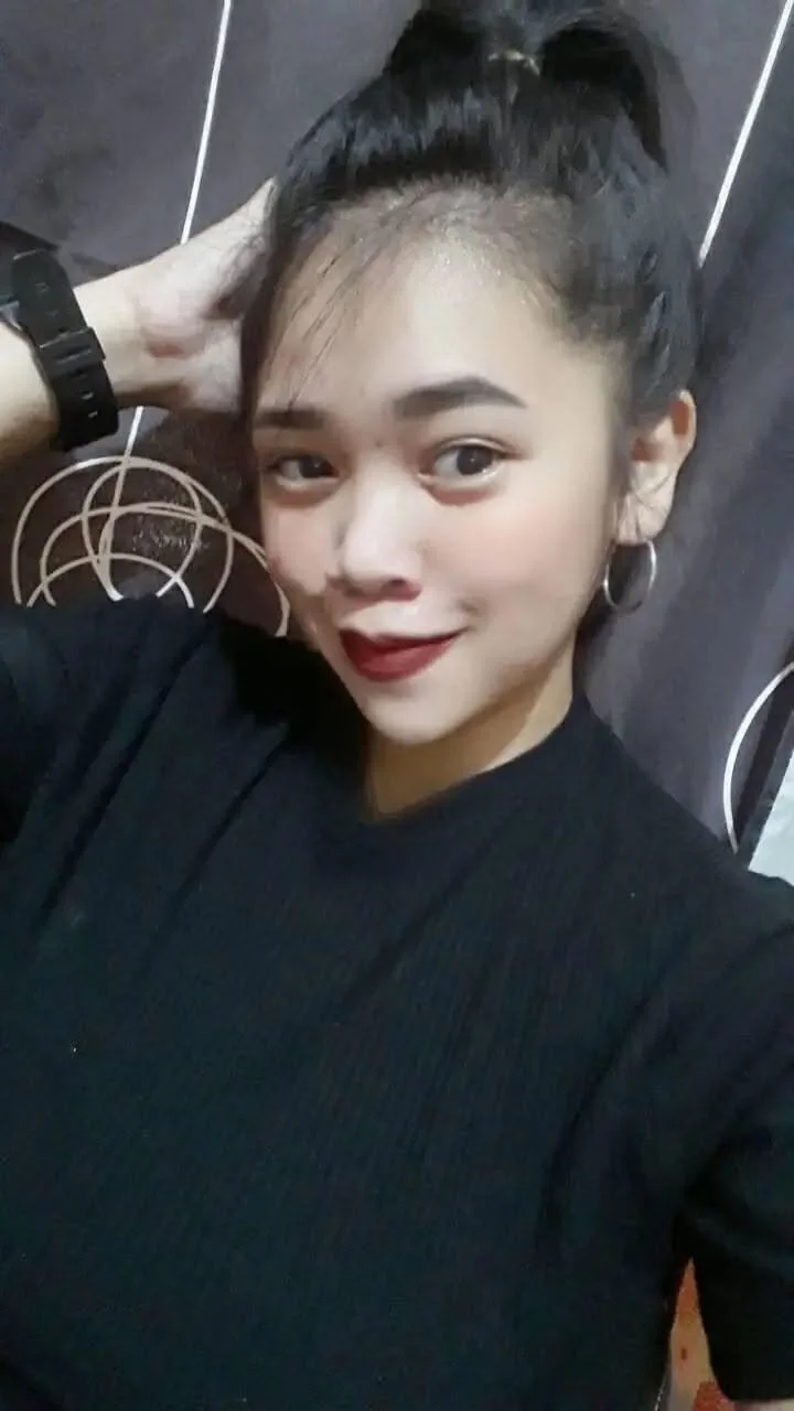 Young woman taking a selfie, smiling, with dark hair tied up in a high ponytail, wearing hoop earrings and a black top, standing against a dark textured background.