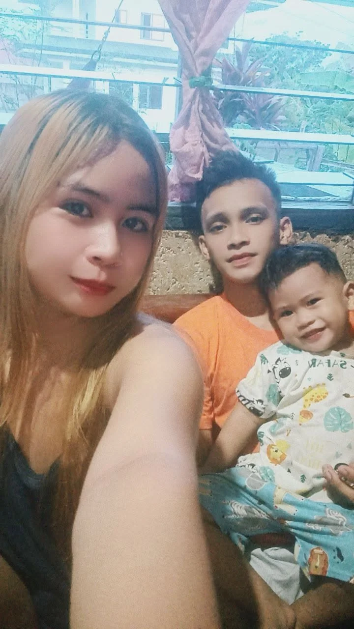 A woman with blonde hair taking a selfie with two young boys, one wearing an orange shirt and the other in pajama with colorful animals, sitting indoors.
