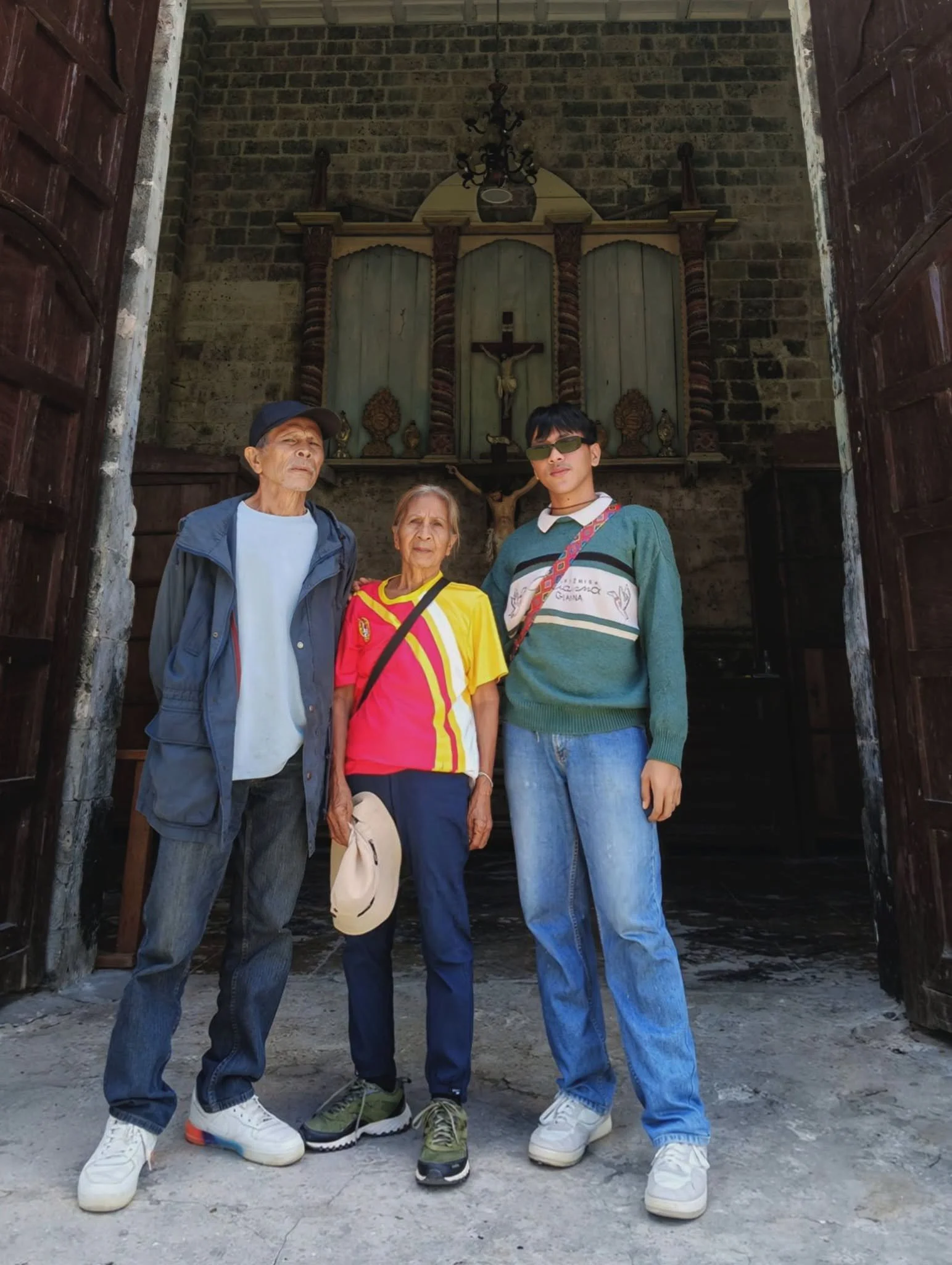 Three people standing together inside a church or chapel with a religious altar behind them. The altar features a crucifix with Jesus Christ on the cross and decorative elements. The individuals are posing for the photo, with the person in the middle