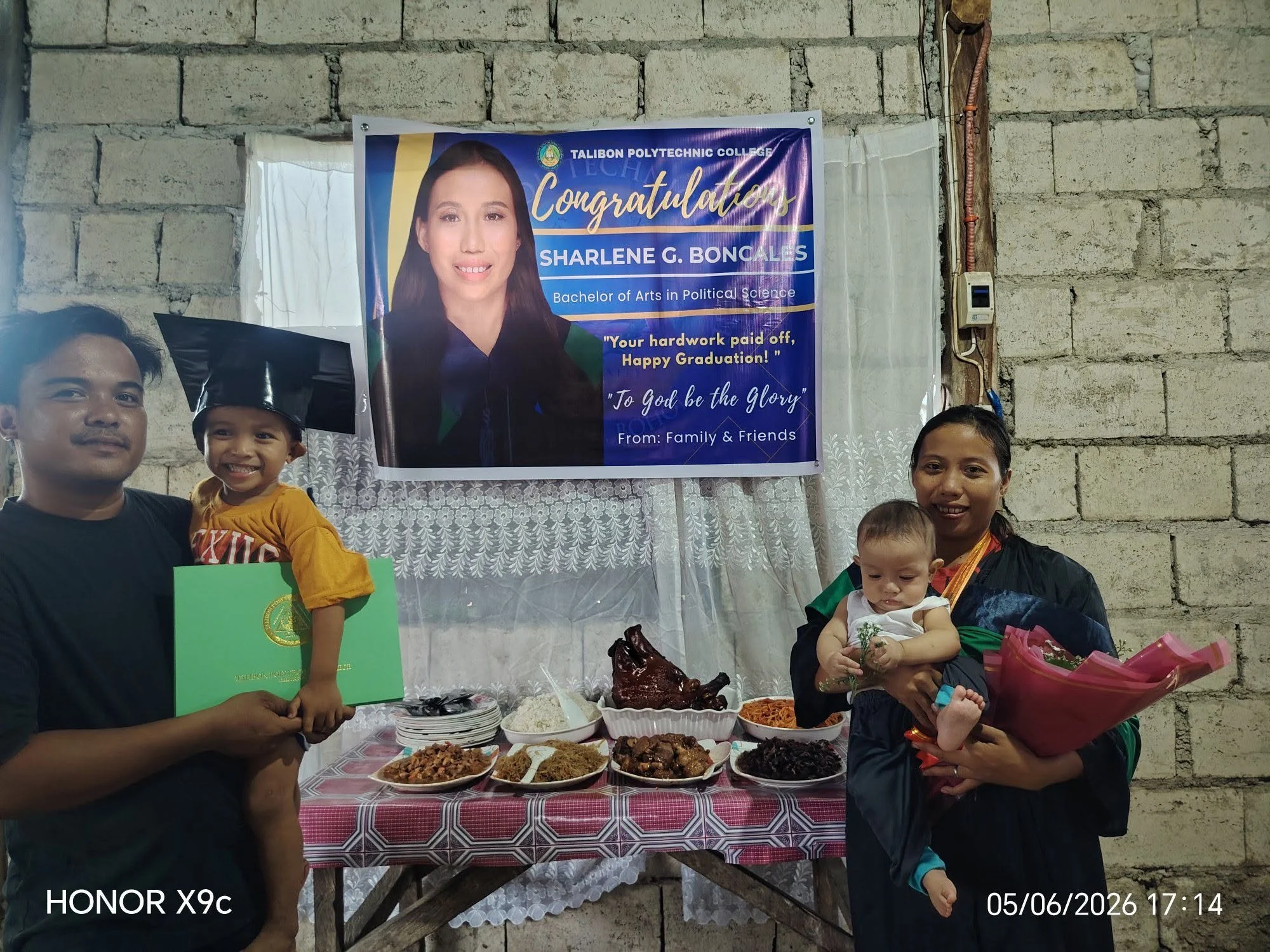 Celebration scene with a family in front of a table filled with traditional dishes. A congratulatory banner for Sharlene G. Boncales, a graduate in Political Science, hangs on a wall behind them. The family members, including young children, are smil