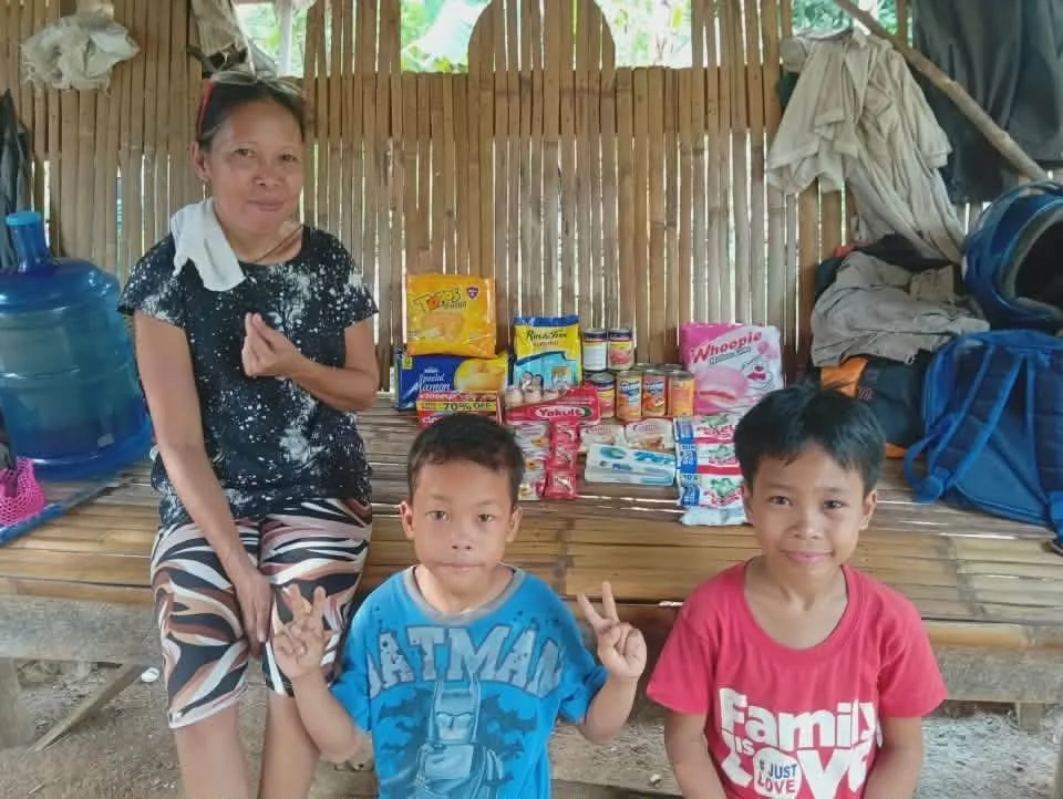 A woman and two young boys sit in front of a display of food and supplies inside a bamboo shelter. The woman is smiling, wearing a black shirt with white spots, and the boys are making peace signs. The supplies behind them include snacks, canned good