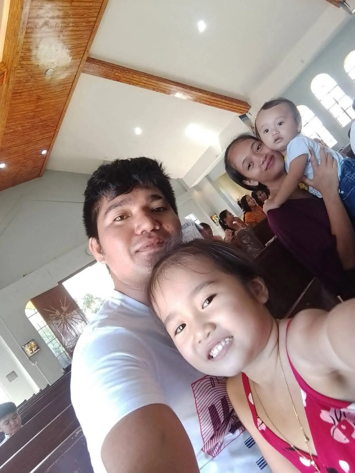A family taking a selfie in a church or community hall, with lights on the ceiling and people sitting in the background.