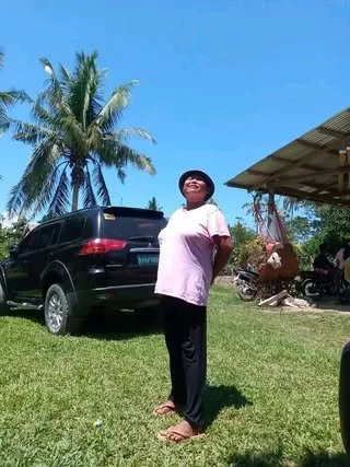 A woman standing outdoors on grass, wearing a pink shirt, black pants, and slippers, smiling and standing near a black SUV, with palm trees and a shelter with motorcycles in the background.