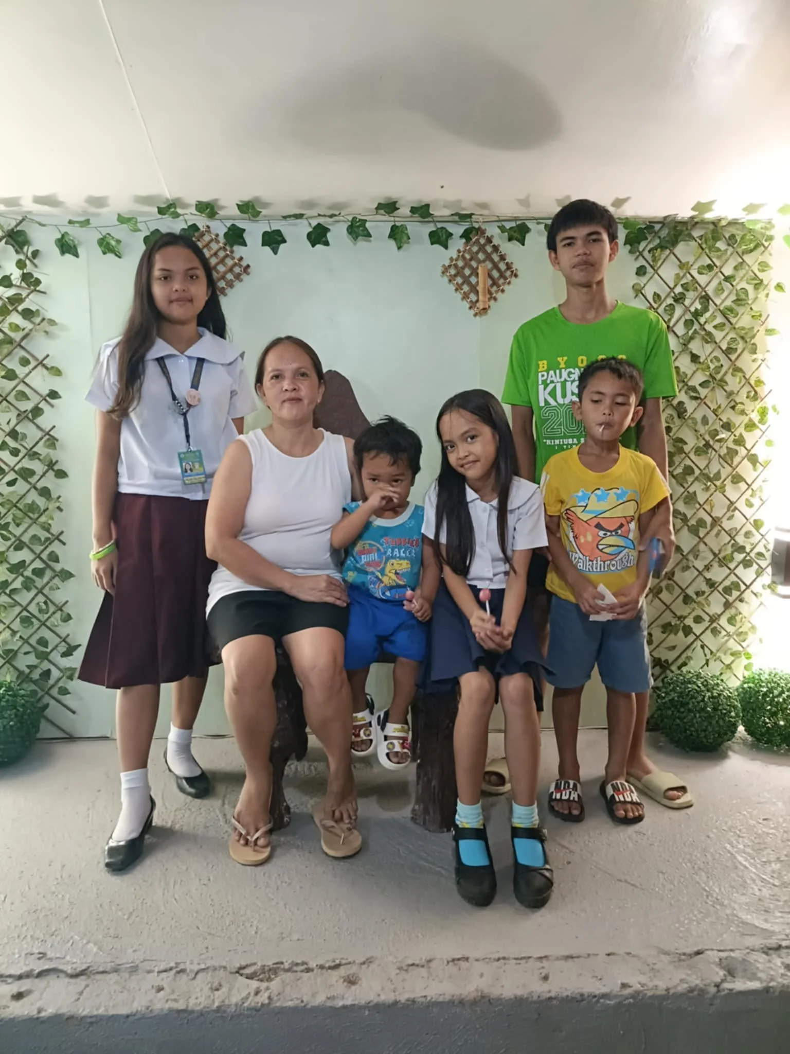 A group of seven people, including children and adults, standing and sitting in front of a decorated wall with green vines and wooden lattice. They are enjoying a moment together indoors, with some smiling and others making different facial expressio