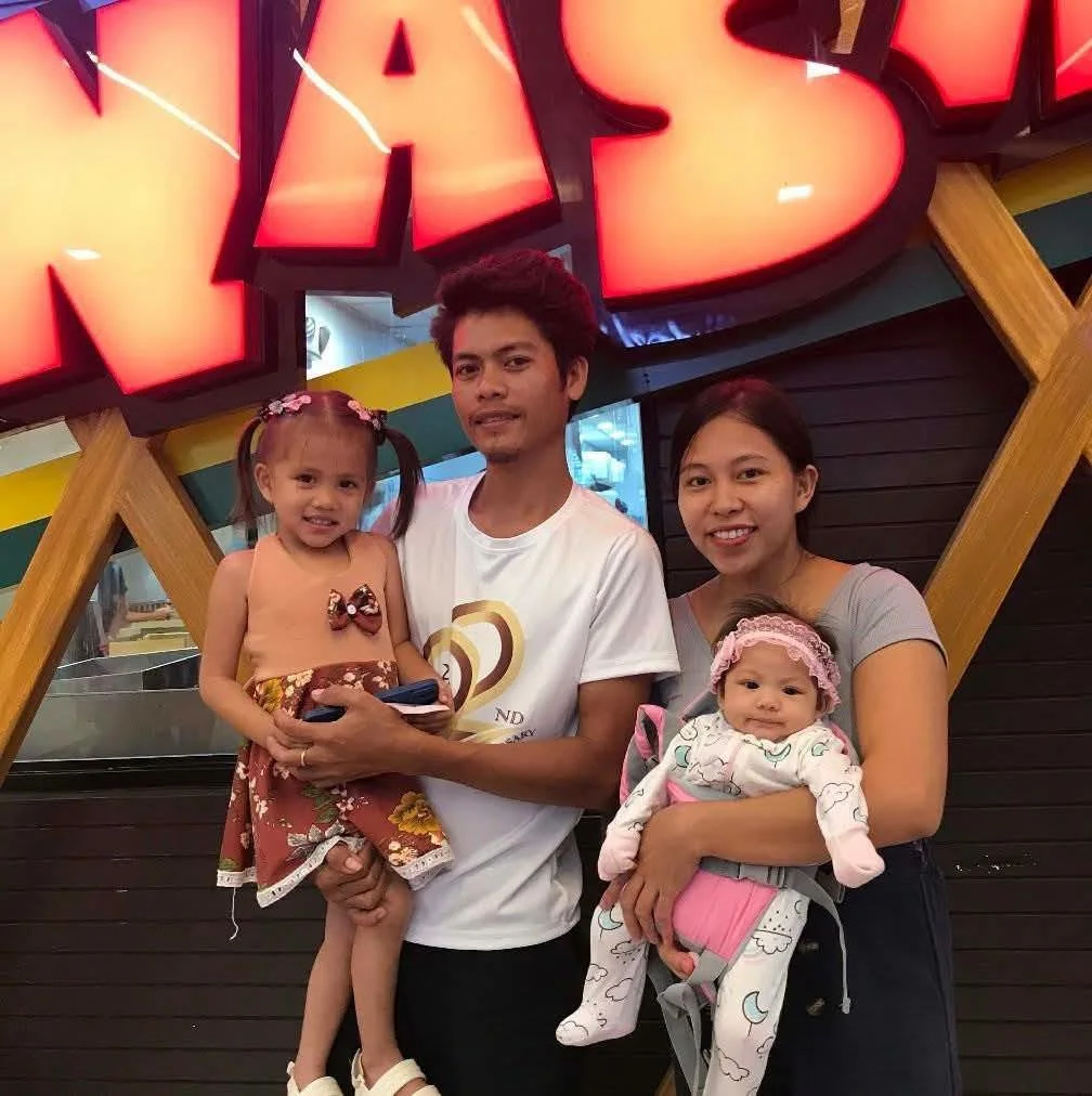 A family of four posing in front of a large sign that reads 'HAPS.' The father is holding a young girl, and the mother is holding a baby girl. The sign is lit up with red and black edges, and the background has wooden and dark paneling.