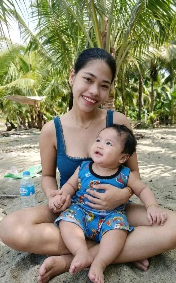 A woman and a young child sitting cross-legged on the sand at a tropical beach with palm trees in the background. The woman is smiling at the camera while holding the child's hand, and the child is looking up at her with a curious expression. The wom