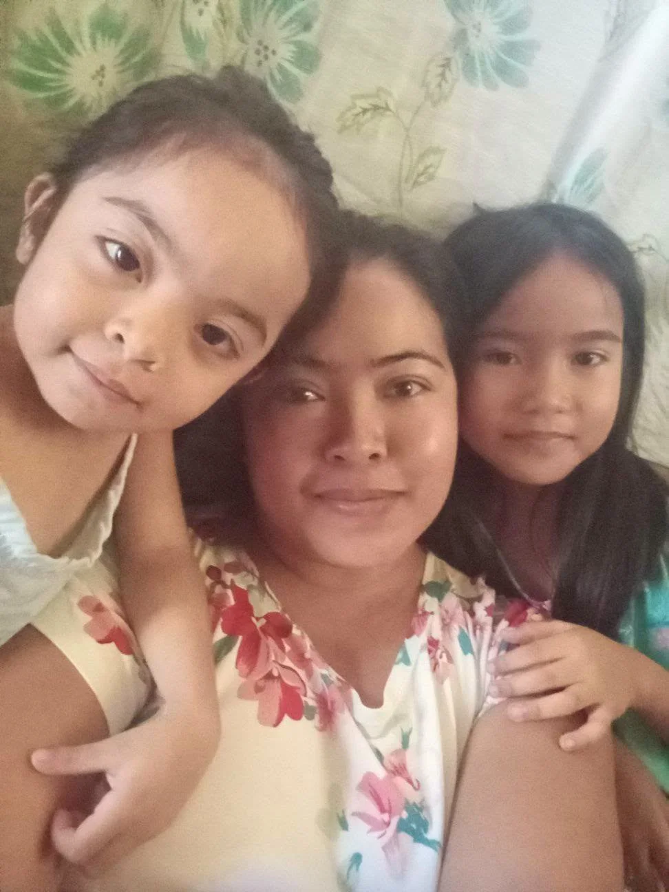 A woman with dark hair and two children, one girl and one boy, lying close together on a bed with a floral bedspread in the background. The woman and girl are smiling softly, while the boy has a curious expression.