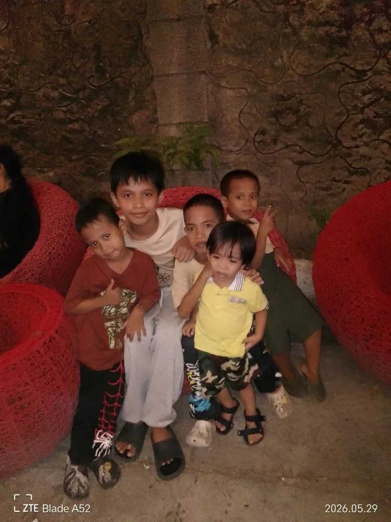 Group of five young children sitting and standing on red woven chairs in front of a stone wall, some smiling and making gestures, in a dimly lit setting.