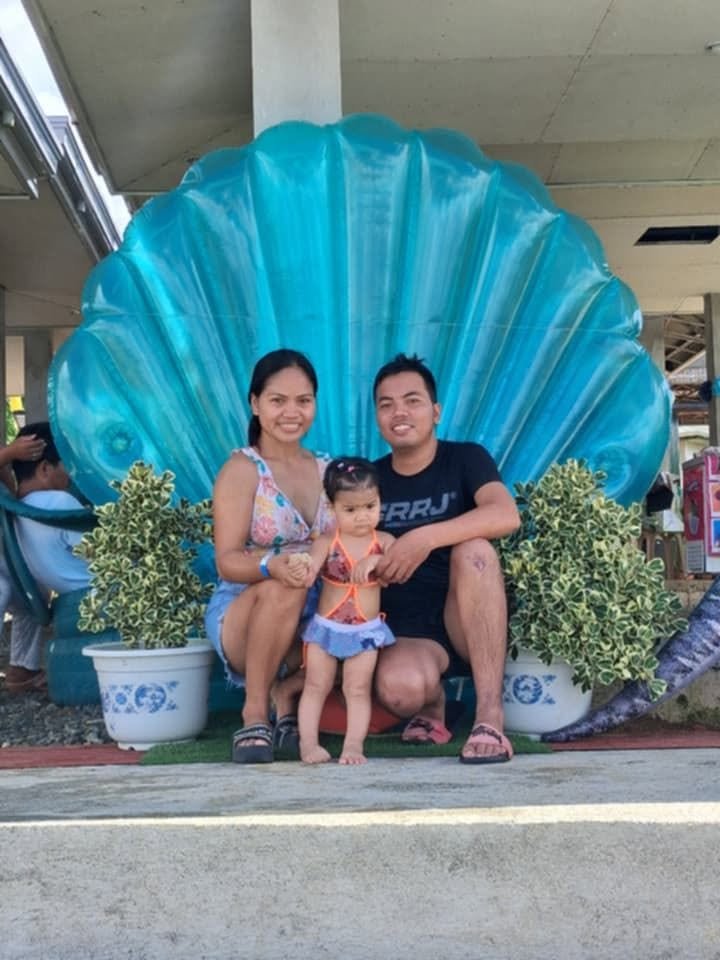 A family of three, a woman, a man, and a young girl, sitting in front of a decorative blue seashell backdrop with potted plants, outdoors at a public place.