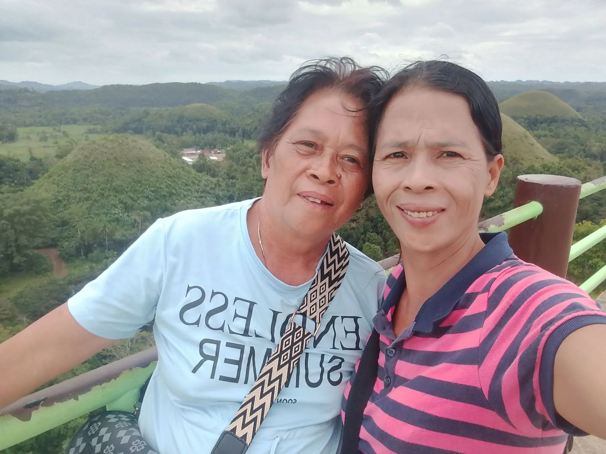 A middle-aged woman and a middle-aged man taking a selfie together outdoors, with green hills and cloudy sky in the background.