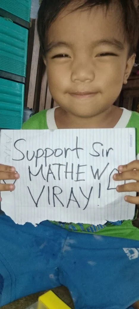 Young boy smiling and holding a sign that says 'Support Sir Mathew Viray'.