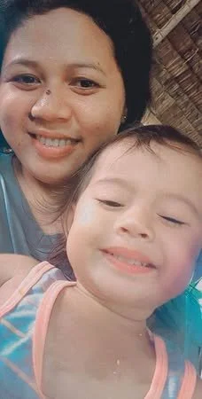A woman with dark hair and a young girl with a smile, both taking a selfie indoors with a wooden wall in the background.