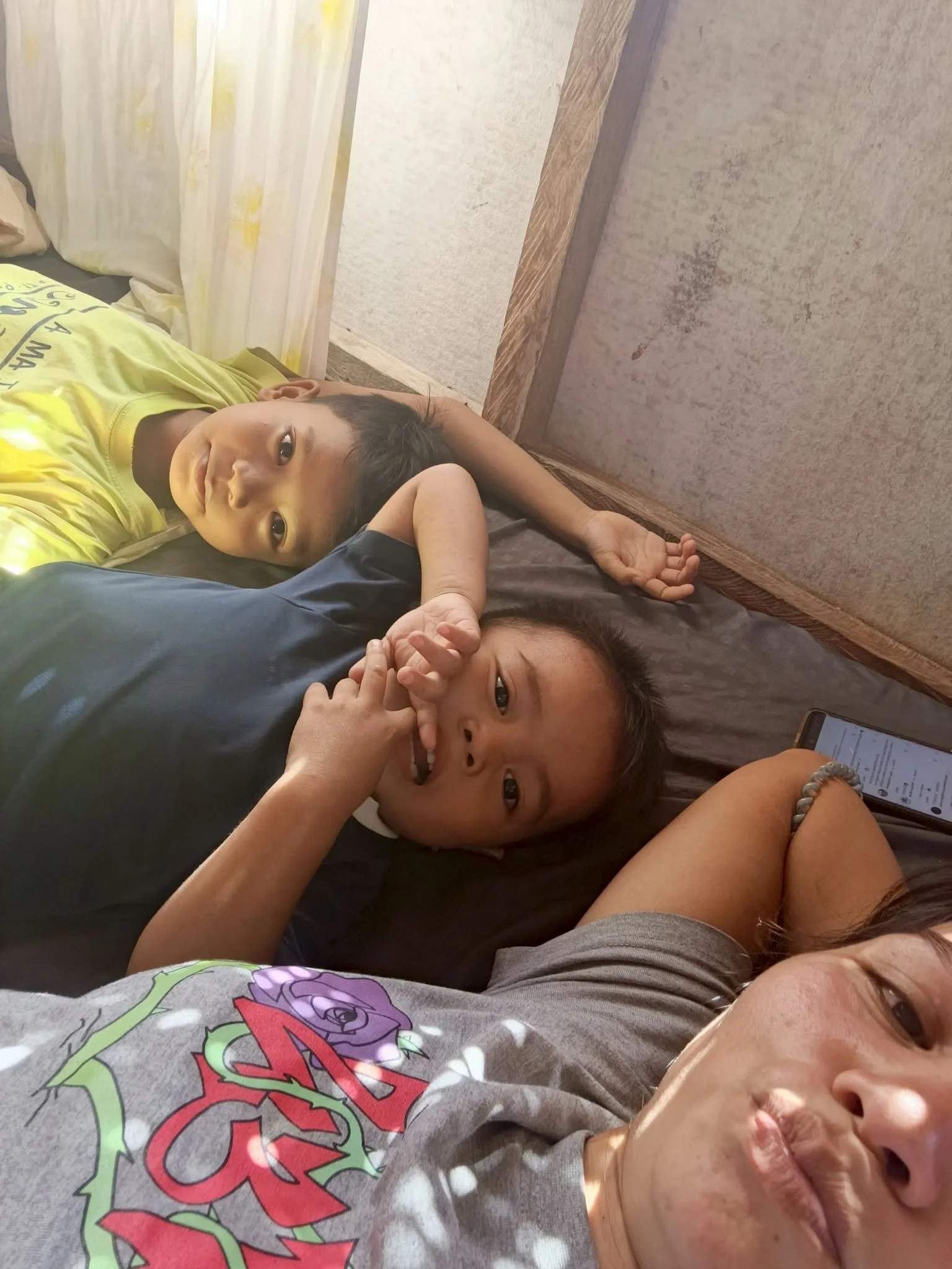 Two children lying on the floor, one with a yellow shirt and the other with a gray shirt and colorful design, and an adult with a watch and a smartphone, near a wall with curtains.