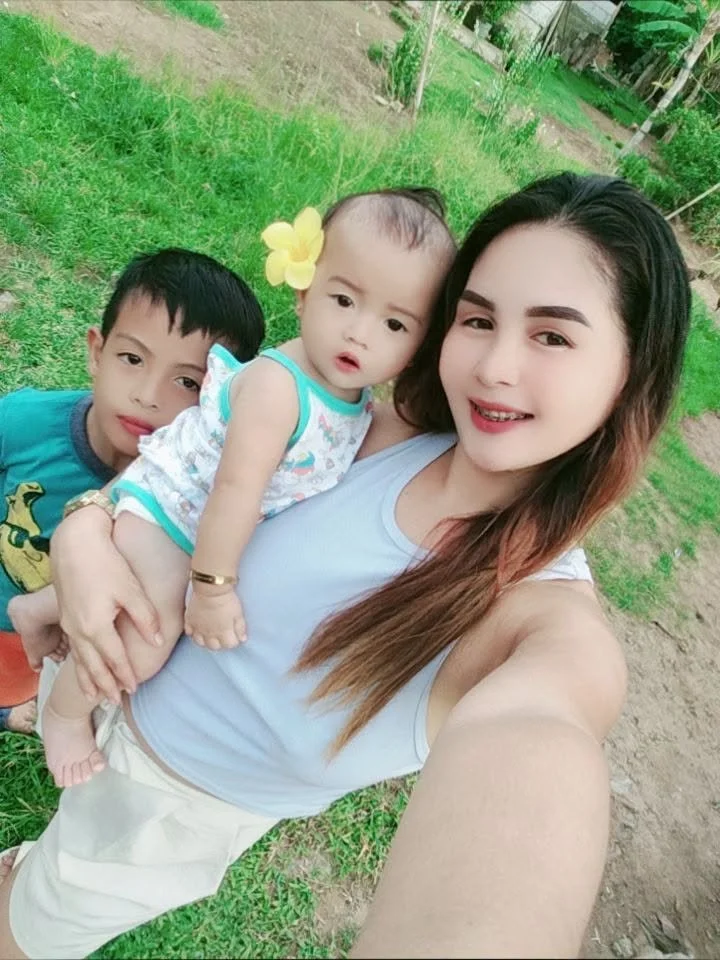 A woman taking a selfie outdoors with two children, one girl and one boy, standing behind her on a grassy area with trees in the background.