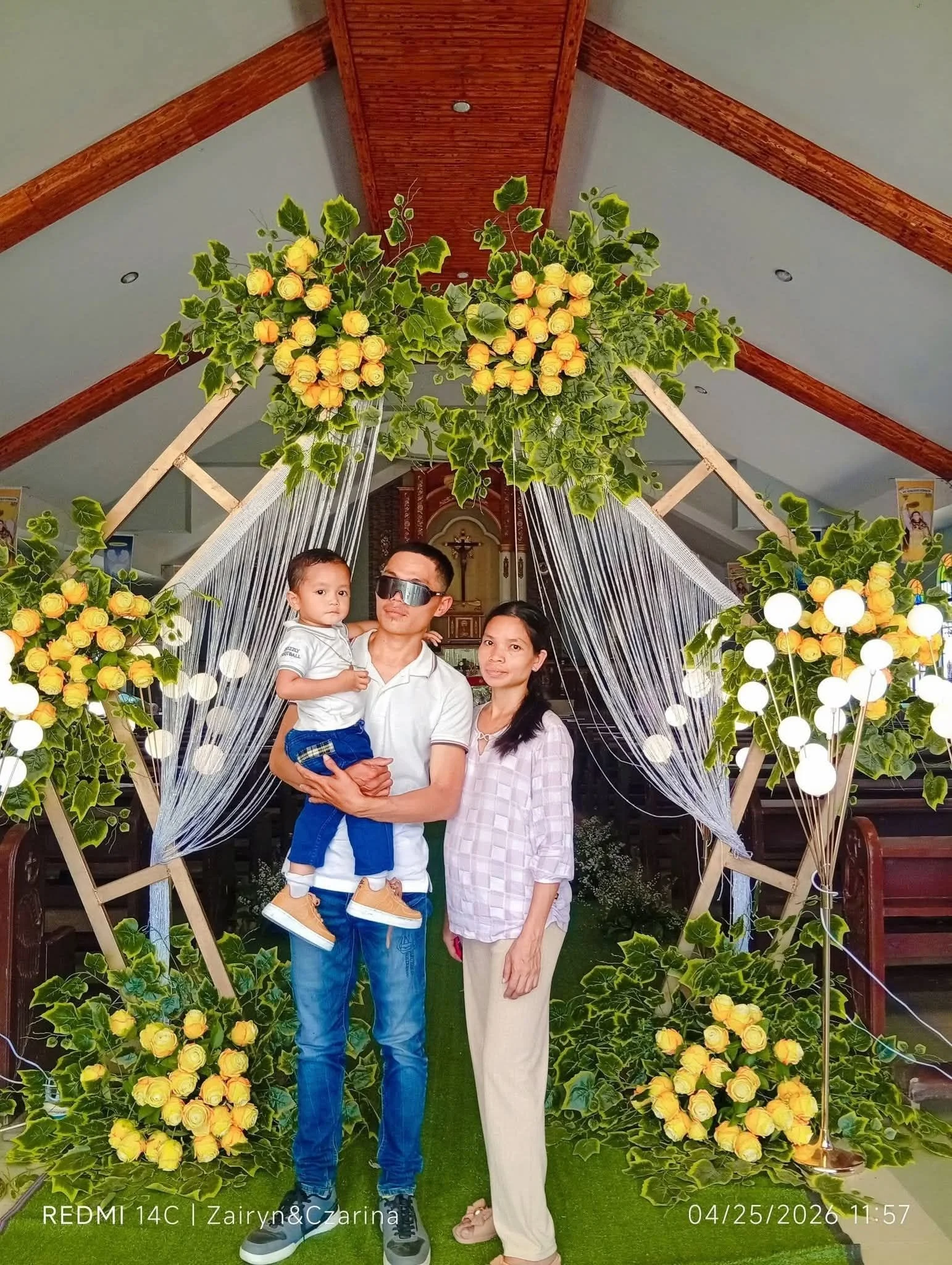 A family of three, including a young boy, a man, and a woman, standing in front of a decorated backdrop with yellow roses, greenery, and decorative lights inside a decorated hall or church. The man is wearing sunglasses, and the woman is dressed in l