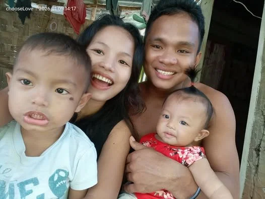 A family of four, including a young boy, a woman, a man, and a baby girl, smiling together outdoors.
