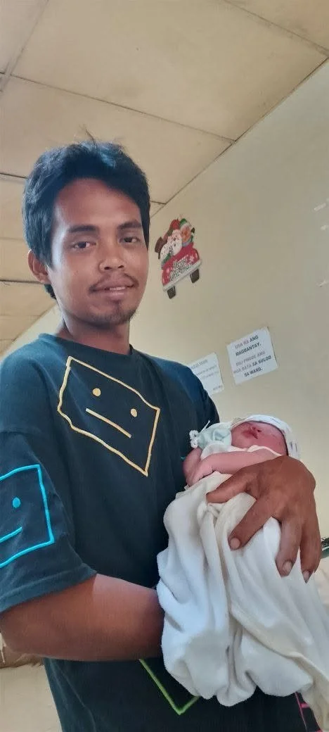 Young man holding a newborn baby wrapped in a white blanket inside a room with beige walls and a ceiling. The man is smiling slightly and is wearing a black shirt with yellow and blue designs.