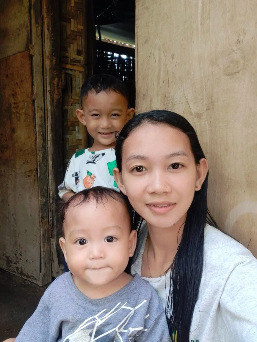 A woman with two young children in front of a wooden wall and doorway, smiling at the camera.