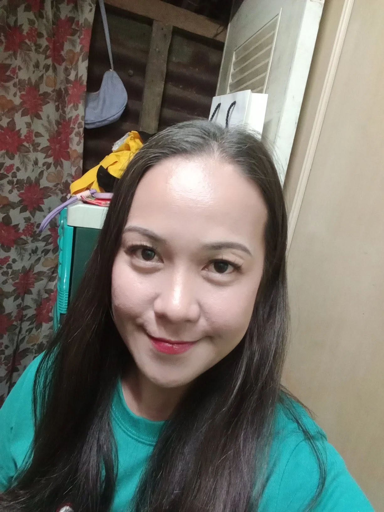 A woman with long brown hair smiling at the camera, wearing a teal shirt, in a room with floral curtains, a plastic container, and a white bag in the background.