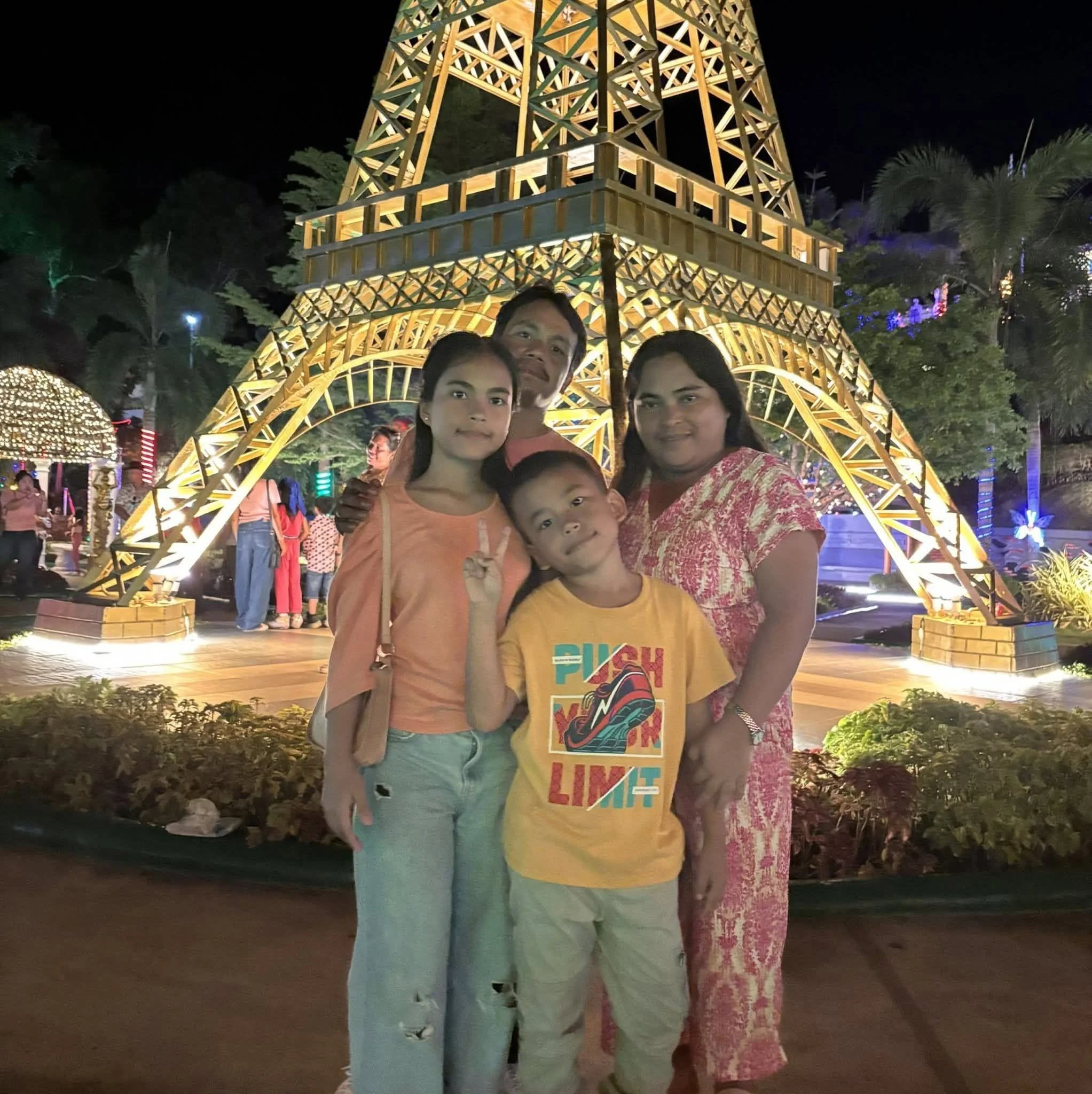 A family of four, including a man, a woman, and two children, standing in front of a large illuminated replica of the Eiffel Tower at night, with trees and other visitors visible in the background.