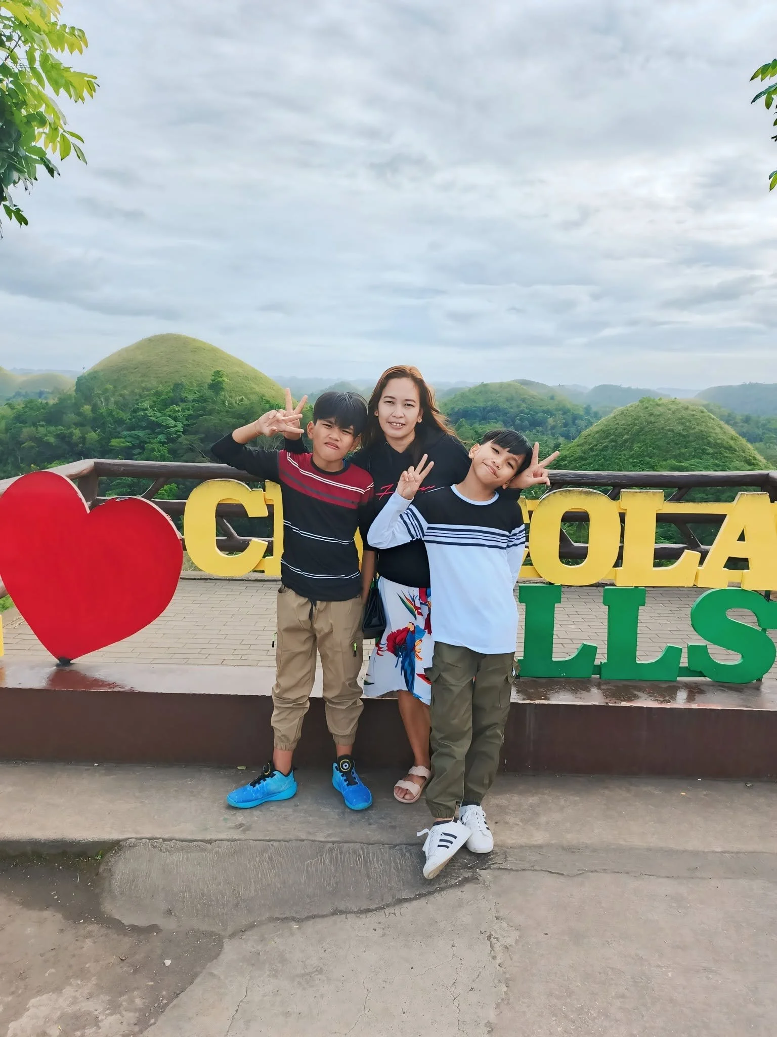 A woman and two young boys posing together outdoors at a scenic overlook with green hills and a cloudy sky in the background. The woman is smiling, making a peace sign with her right hand, while the boys are also making peace signs.