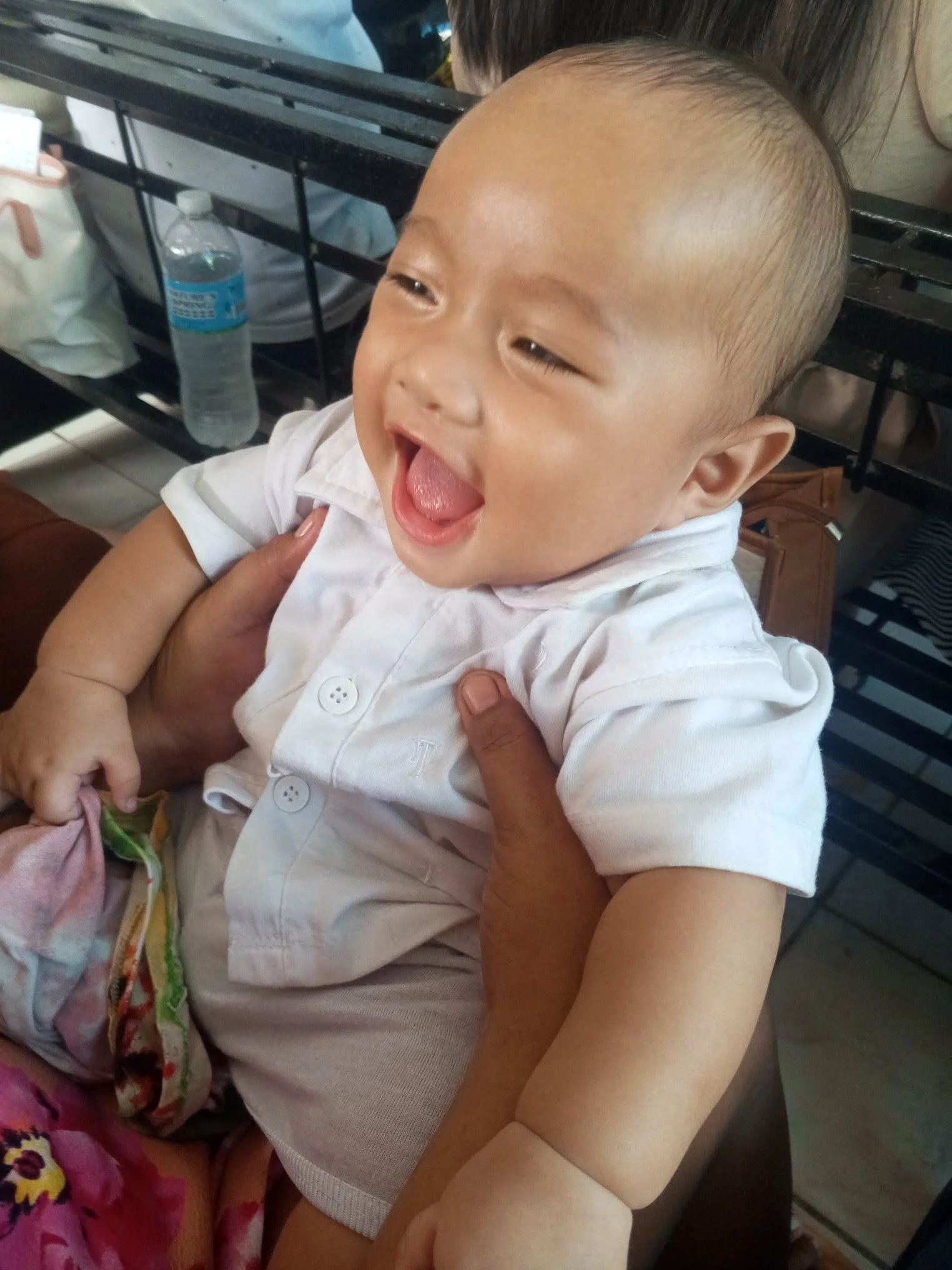 A young boy with a shaved head, wearing a white shirt, is laughing and being held by an adult. There is a water bottle and bags on a black metal rack in the background.