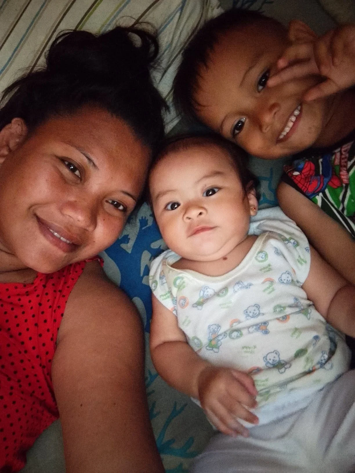 A woman and two children lying on a bed, smiling at the camera. The woman is wearing a red, polka-dot top, and the children are smiling with one showing a peace sign. The baby is in a light-colored onesie with cartoon animals.