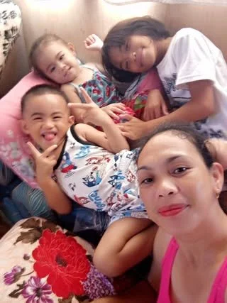 Four children lying on a bed, smiling and making peace signs. They are in a cozy room with floral blankets and pillows.