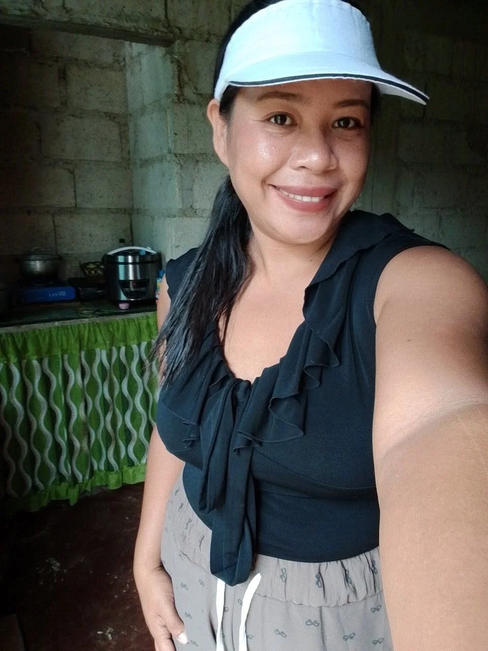 A woman smiling, wearing a white visor and black top, taking a selfie indoors with a concrete wall and kitchen area in the background.
