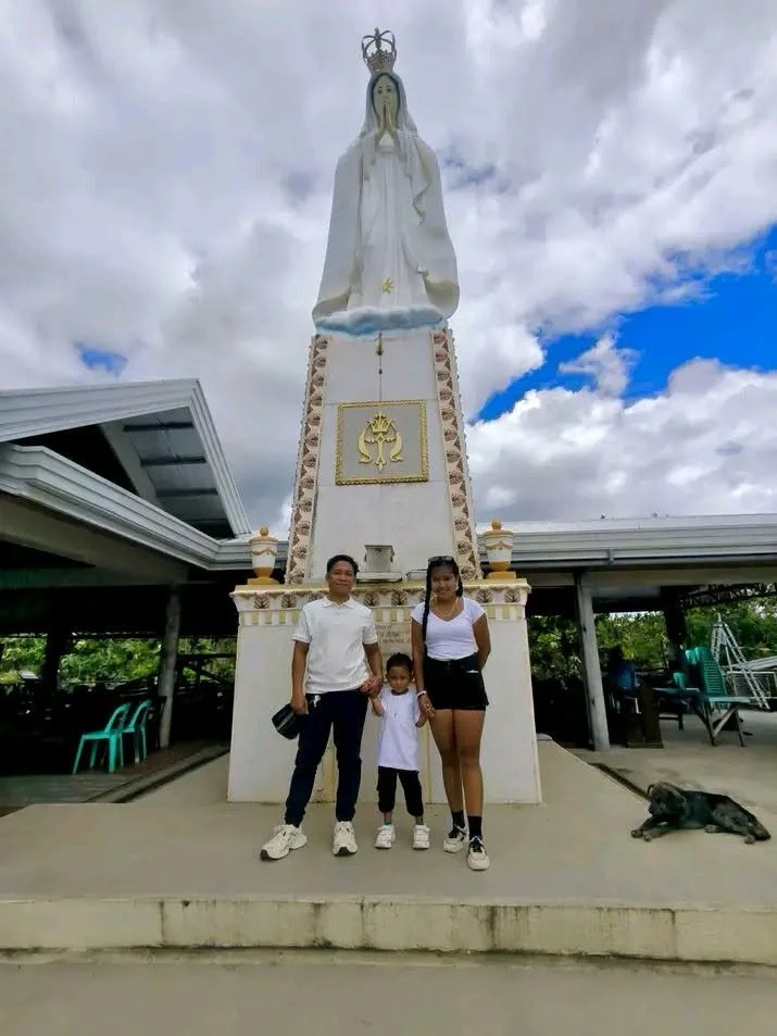 Family standing in front of a large statue of the Virgin Mary outdoors, with cloudy sky in the background.