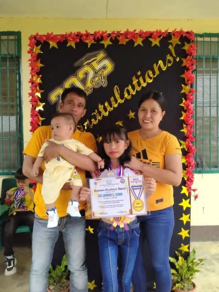 Family celebrating a graduation with a young girl holding a certificate and medal, flanked by a man and a woman, in front of a decorated black backdrop with yellow stars and the word 'Congratulations.'