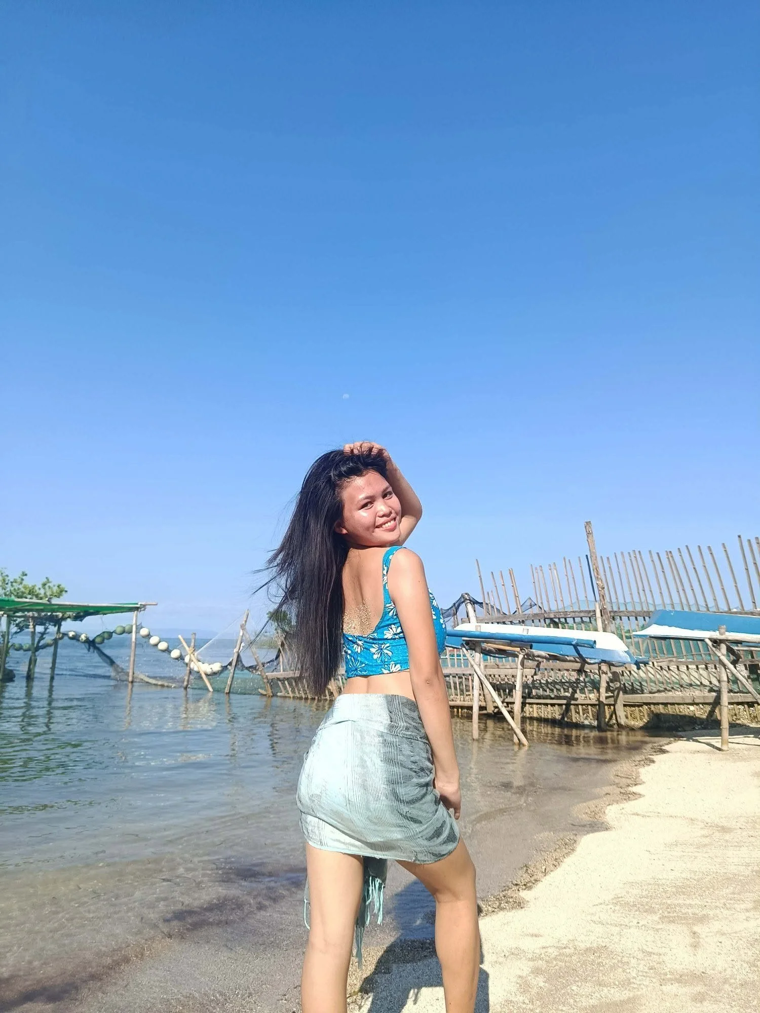 A young woman at the beach posing with her right hand on her head and smiling. She has long dark hair and is wearing a blue floral top and gray shorts. In the background, there are wooden structures and a cloudy sky.