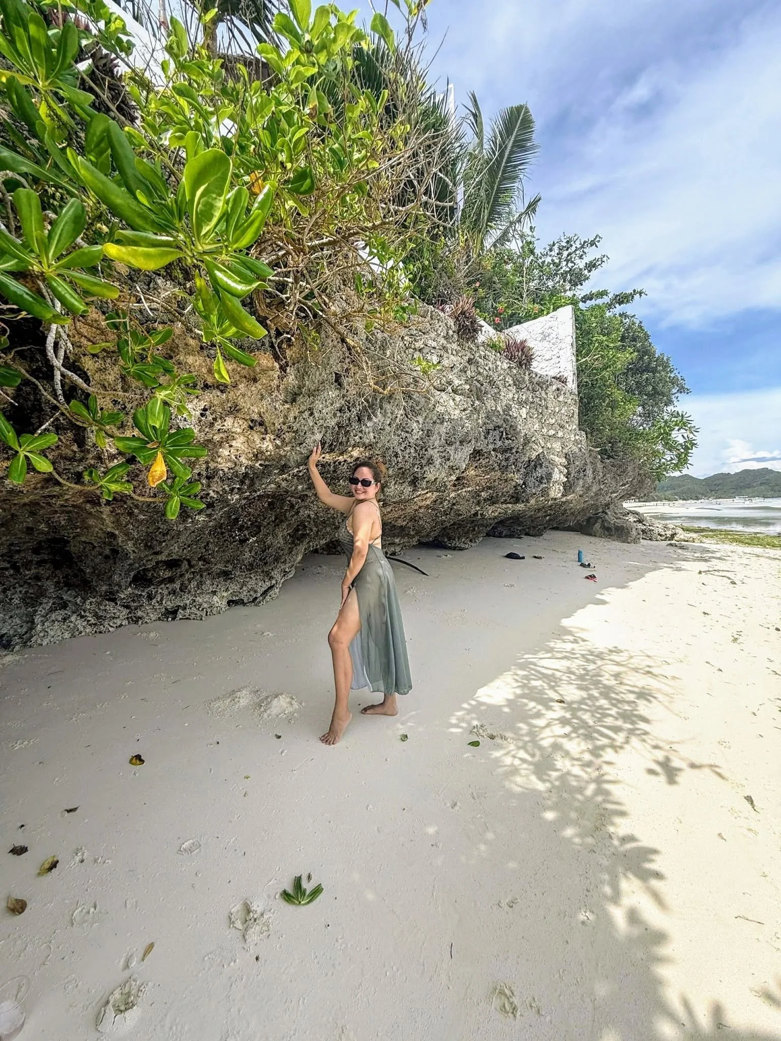 A woman standing on a sandy beach under a large rock formation with greenery on top, smiling and wearing sunglasses and a casual skirt. The sky is partly cloudy and the ocean is visible in the background.