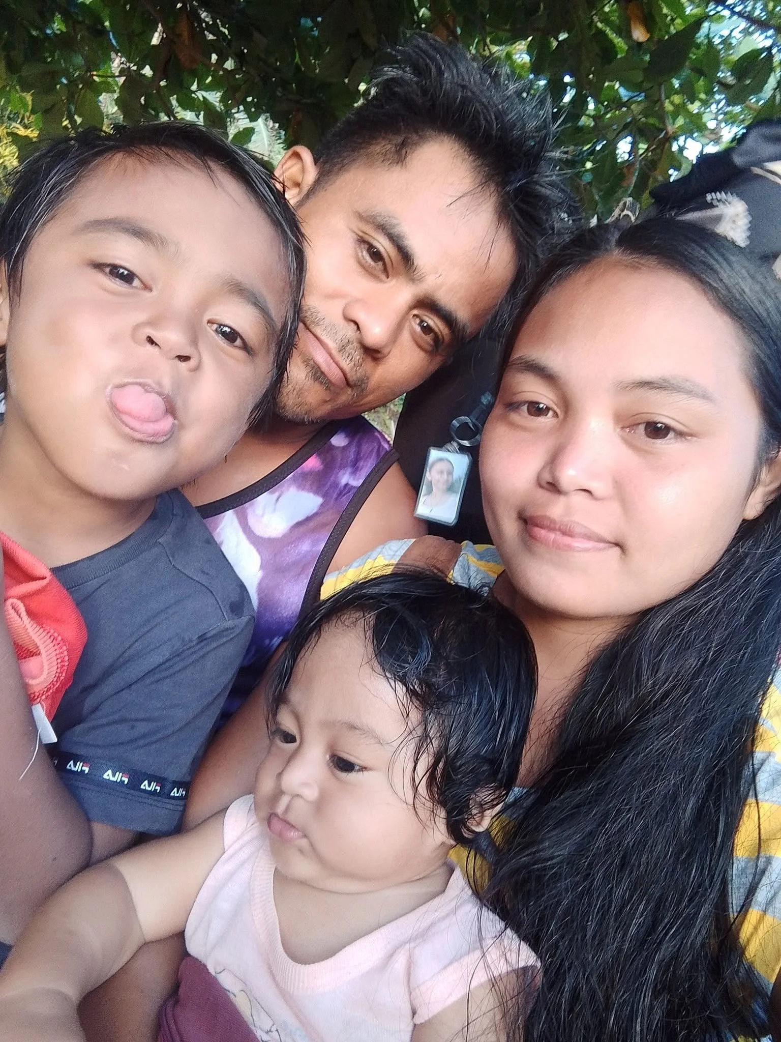A close-up selfie of a family with a man, a teenage girl, a young boy making a silly face, and a young girl, outdoors surrounded by green leaves.
