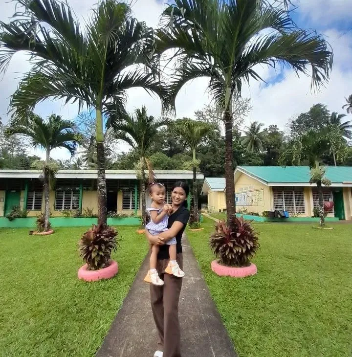A woman holding a young girl on a pathway in a grassy area with palm trees, trees, and buildings in the background.