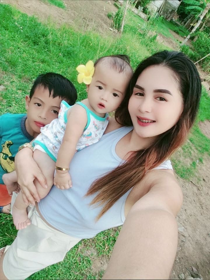 A woman taking a selfie outdoors with two children, one girl and one boy, standing on a grassy area with dirt patches and green trees in the background.